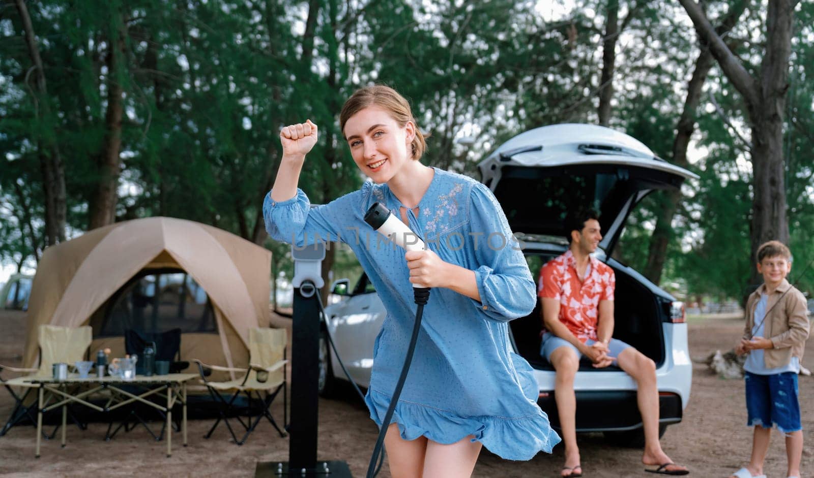 Outdoor adventure and family vacation camping at sea travel by eco friendly car. Cheerful woman or mother holding, pointing EV charger point with playful and happiness posture in campsite. Perpetual