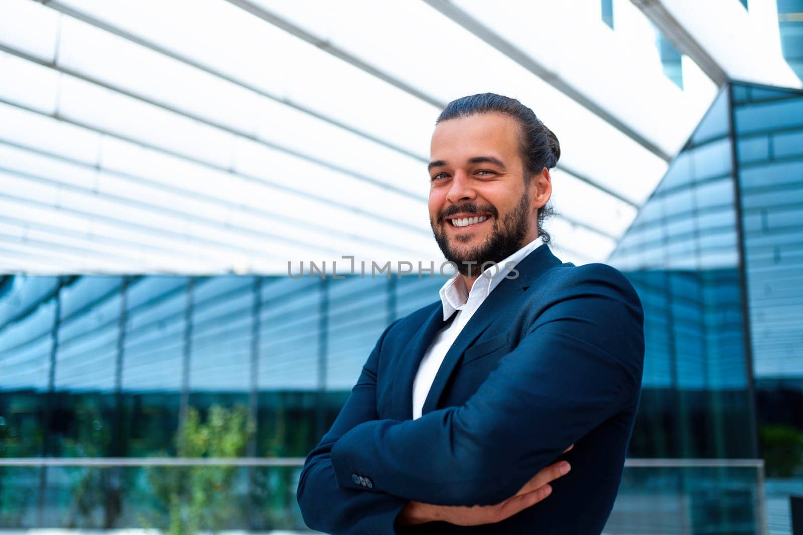 Confidence successful businessman or worker in suit with beard standing in front office glass building arm crossed looking at camera and smile. Hispanic male business person side view portrait.
