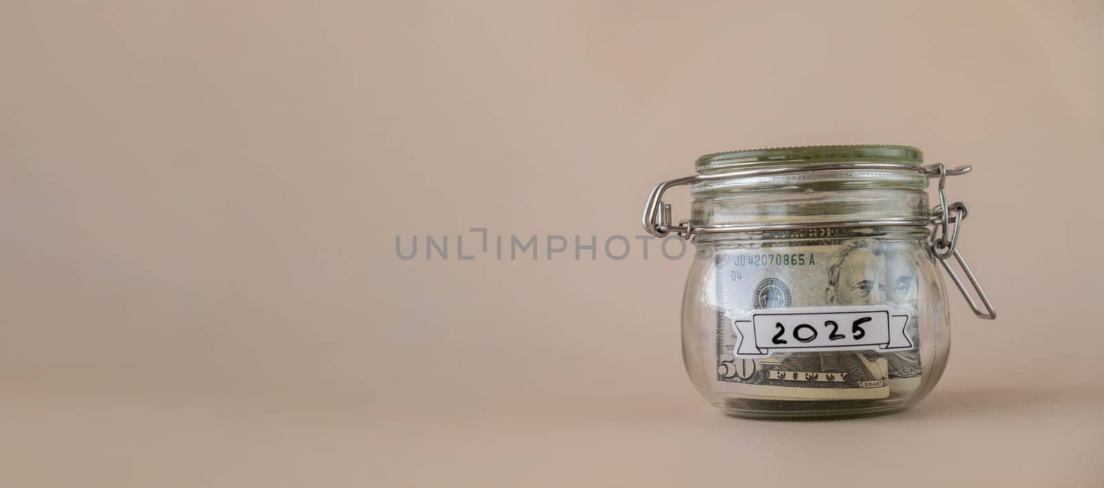 Saving Money In Glass Jar filled with Dollars banknotes. 2025 year transcription in front of jar. Managing personal finances extra income for future insecurity by anna_stasiia