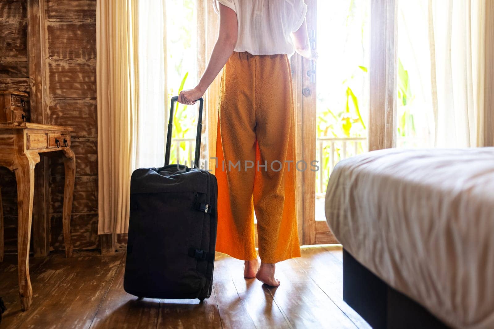 Rear view of unrecognizable woman walking out of hotel bedroom with suitcase. by Hoverstock