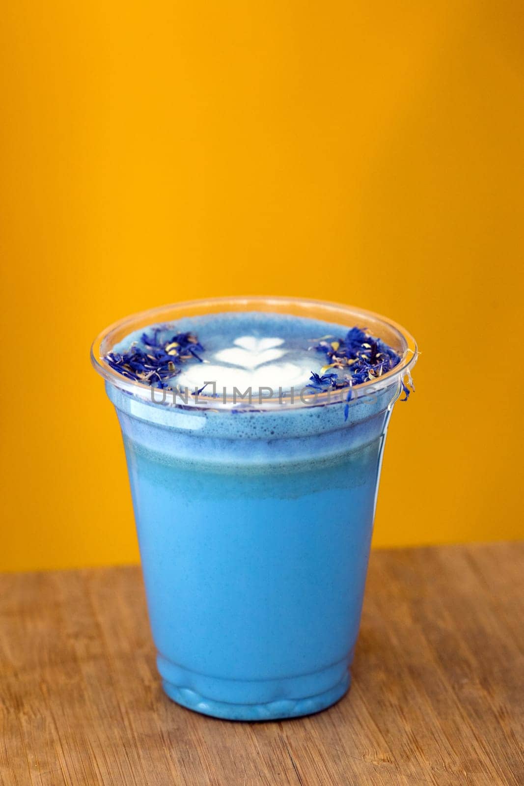 Blue drink matcha milk tea with flowers and foam with a heart pattern on a yellow background in a plastic glass to take away
