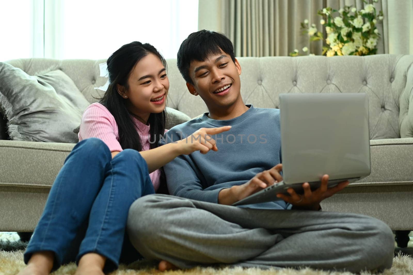 Happy young couple in casual outfit sitting carpet in living room and using laptop together by prathanchorruangsak