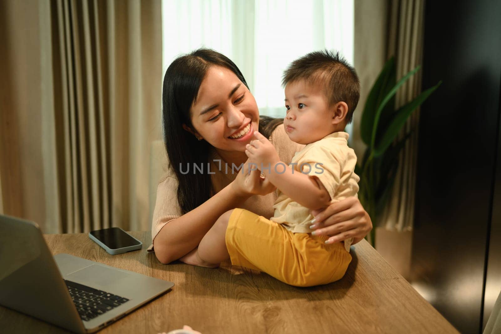 Working mom working online on laptop and taking care of her baby at kitchen table by prathanchorruangsak