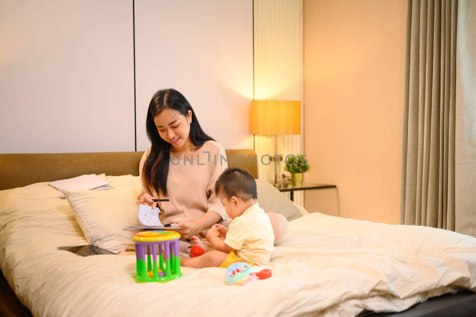 A cute baby boy playing toys in comfortable bed near working mother. Childcare and working mom concept.