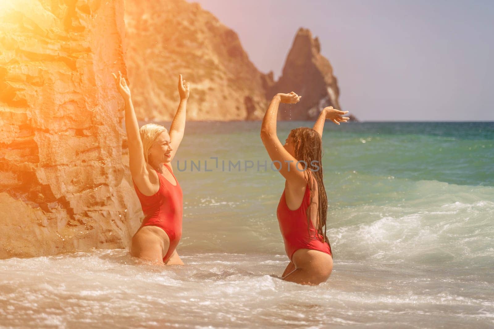 Women Santa hats ocean play. Seaside, beach daytime, enjoying beach fun. Two women in red swimsuits and Santa hats are enjoying themselves in the ocean waves and raising their hands up. by Matiunina