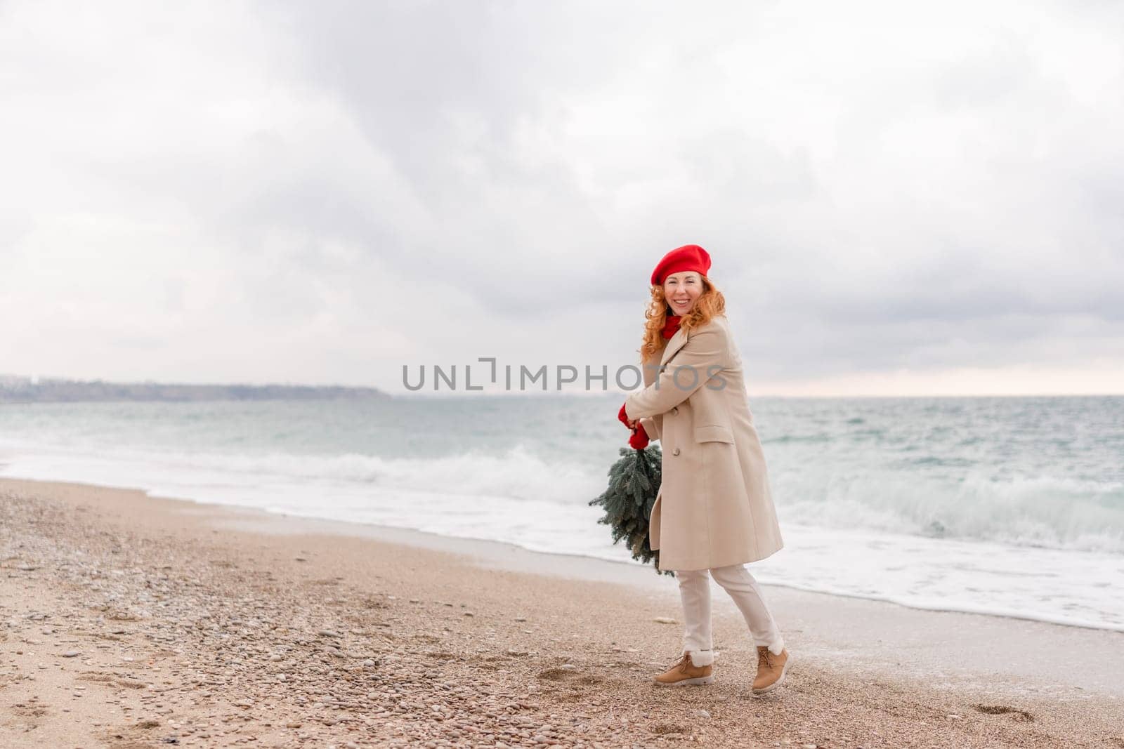 Redhead woman Christmas tree sea. Christmas portrait of a happy redhead woman walking along the beach and holding a Christmas tree in her hands. She is dressed in a light coat and a red beret