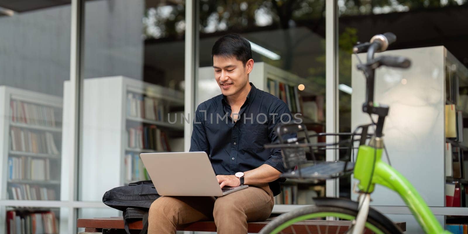 Asian businessman with bicycle using laptop and sitting outside the office building. man commuting on bike go to work. Eco friendly vehicle, sustainable lifestyle concept.