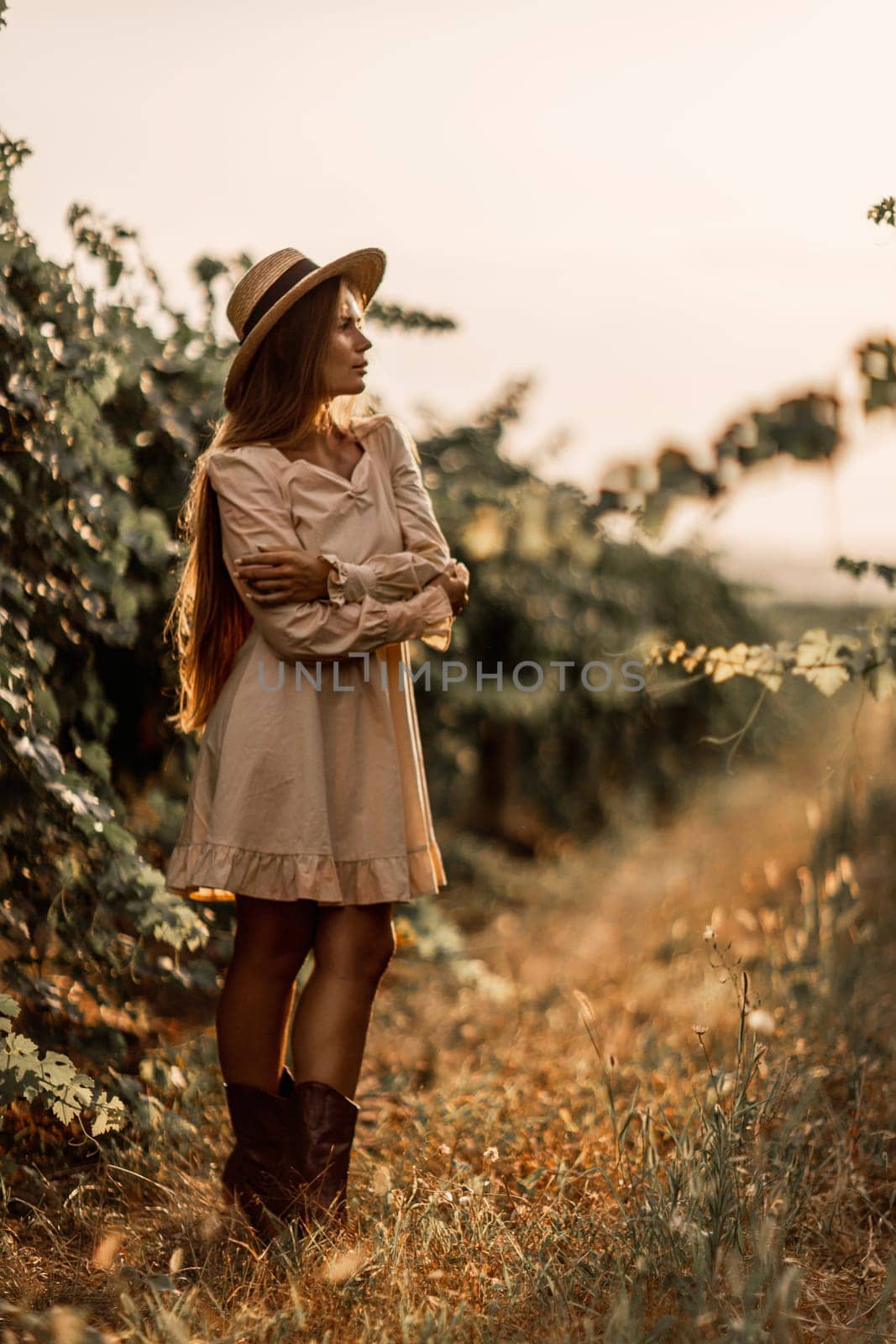 Woman with straw hat stands in front of vineyard. She is wearing a light dress and posing for a photo. Travel concept to different countries.