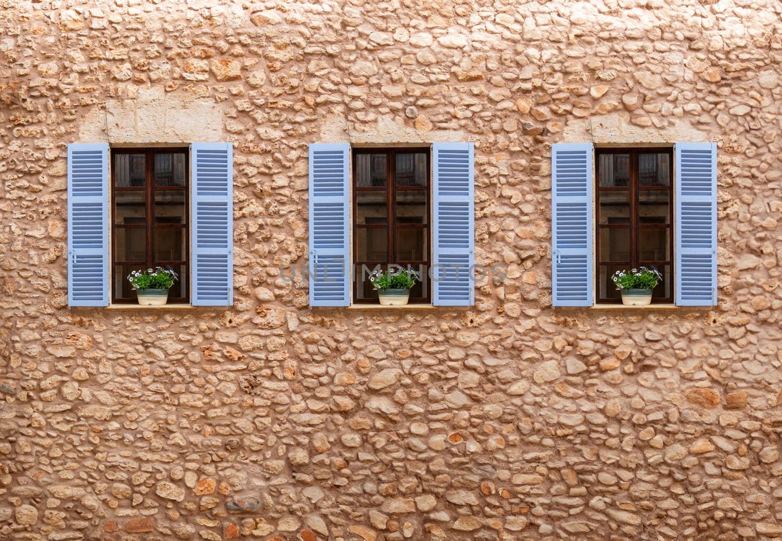 Three windows with blue shutters and white flower boxes on a rustic stone wall.