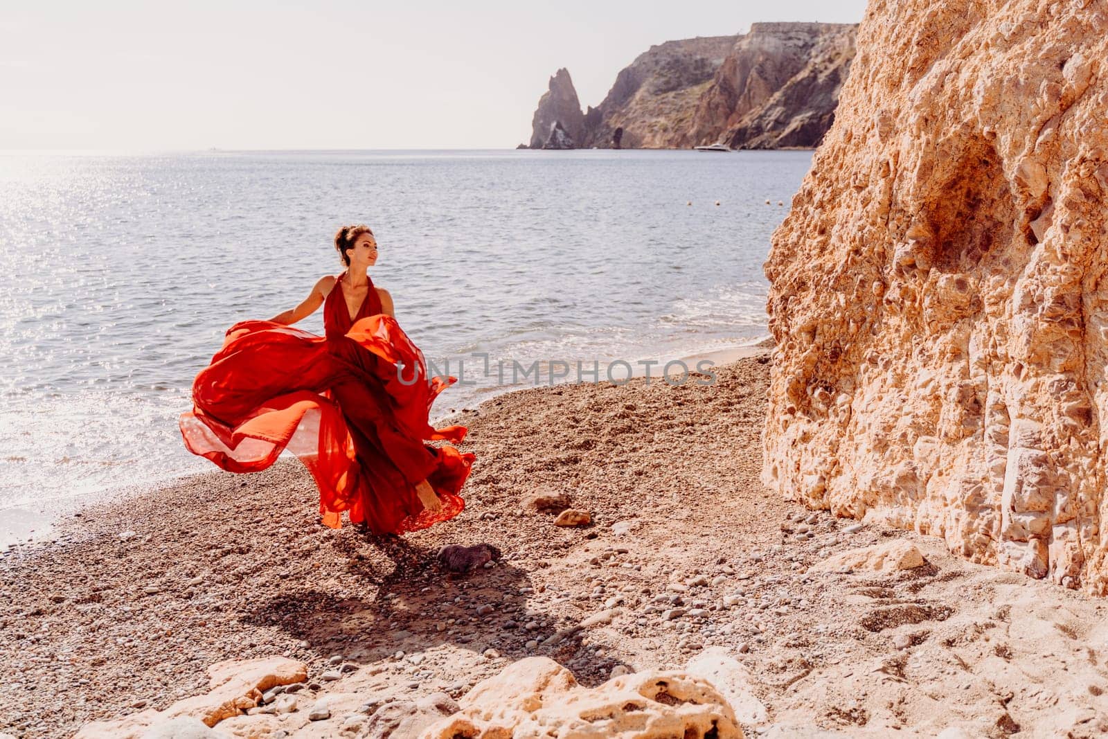 Woman red dress sea. Female dancer in a long red dress posing on a beach with rocks on sunny day. Girl on the nature on blue sky background