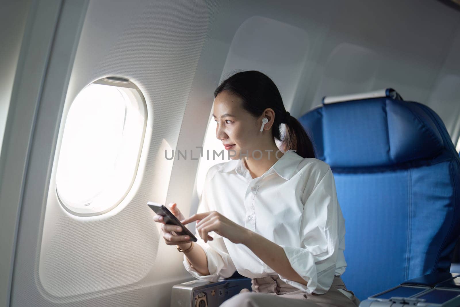A woman is sitting on an airplane with her phone in her hand. She is looking at the screen and pointing at something