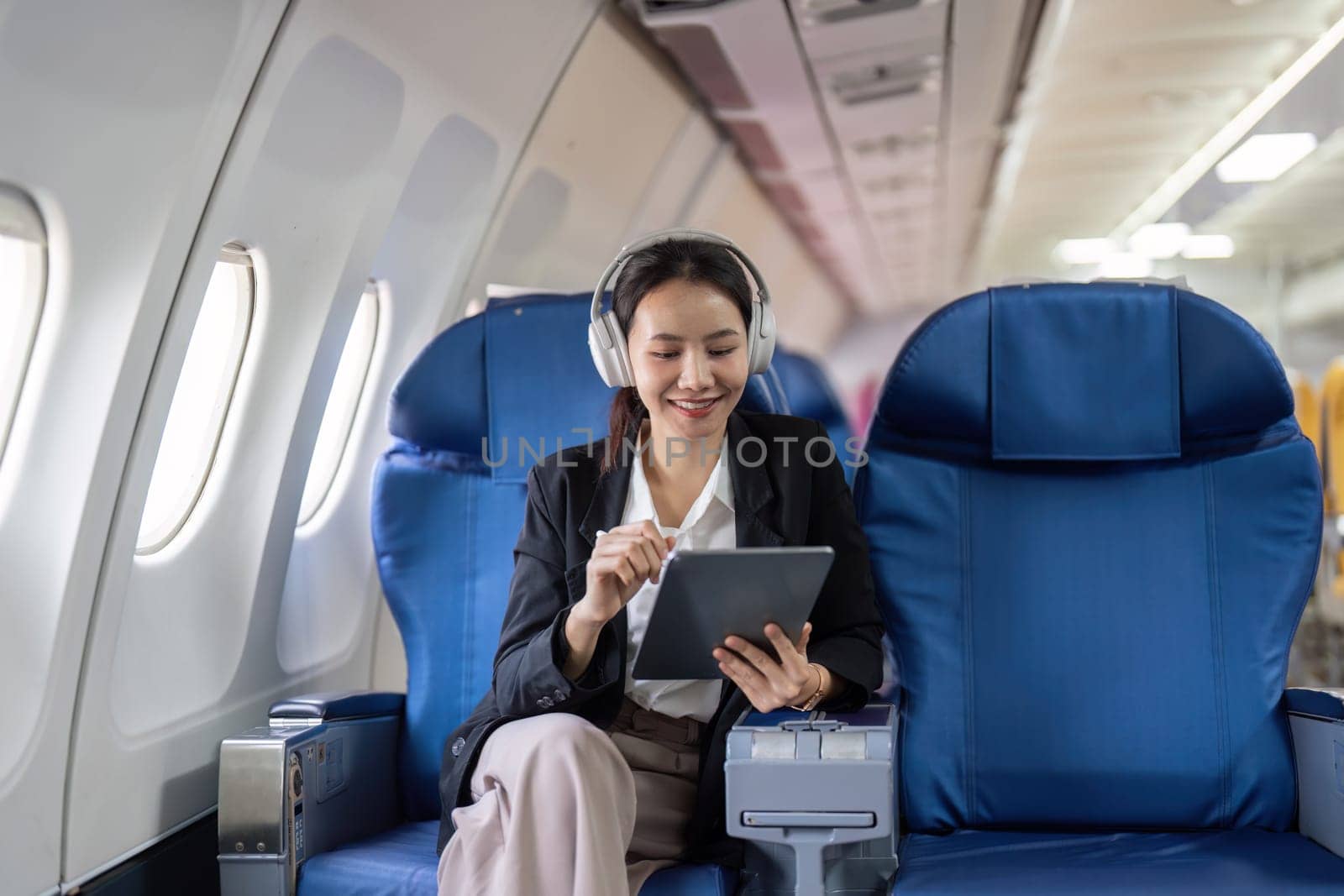 A woman is sitting on a blue airplane seat with a tablet in her hand. She is smiling and she is enjoying her time on the plane