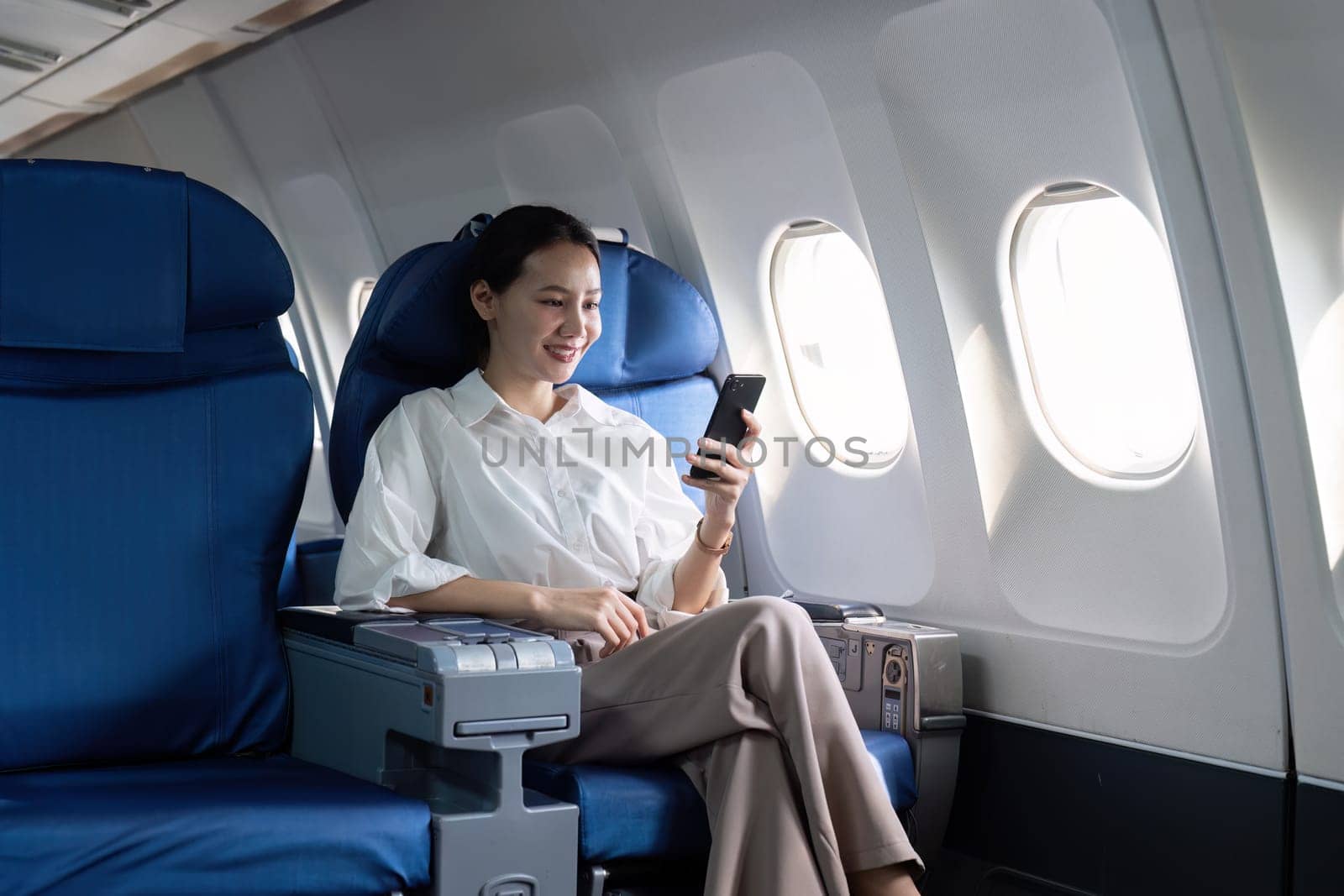 A woman is sitting in an airplane seat with a cell phone in her hand. She is smiling and she is enjoying her time on the plane