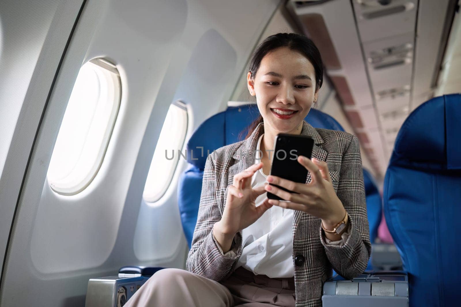 Young Asian woman checks business news on mobile phone, sitting near window in first class on airplane during flight, travel and business concept.