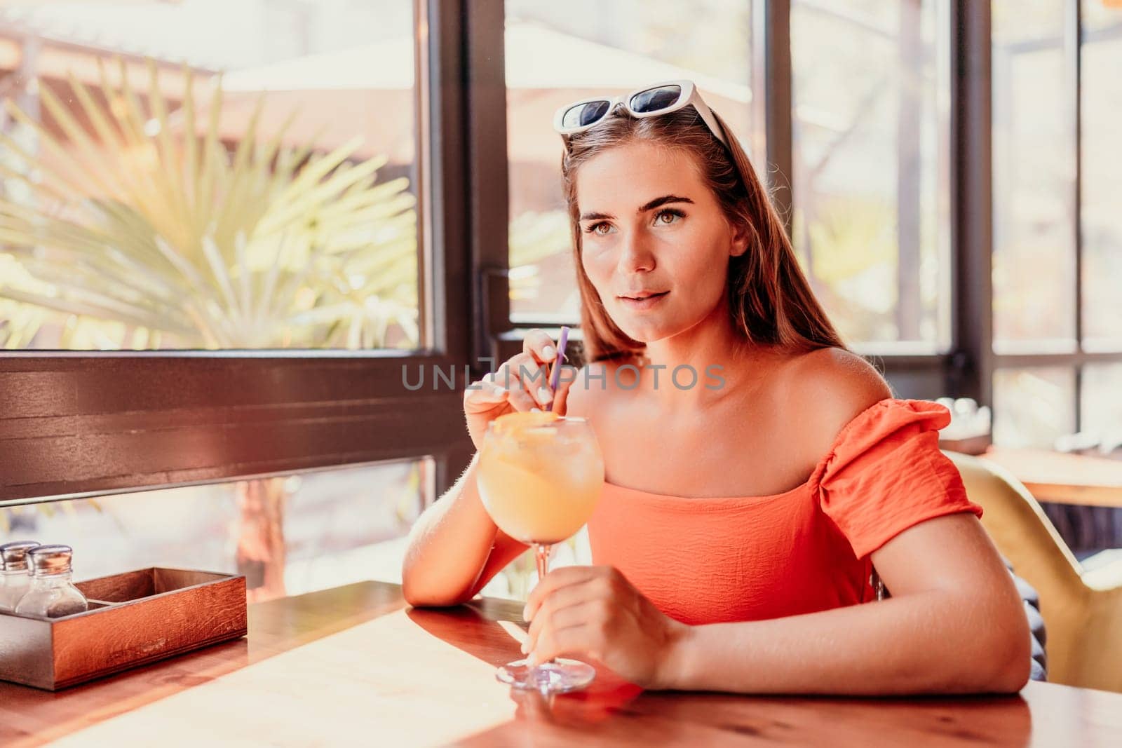 woman in a cafe seated, holding beverage. Dressed in an orange dress with her hair down. Bright interior, large windows allow natural light. Beverage consumption for refreshment. by Matiunina