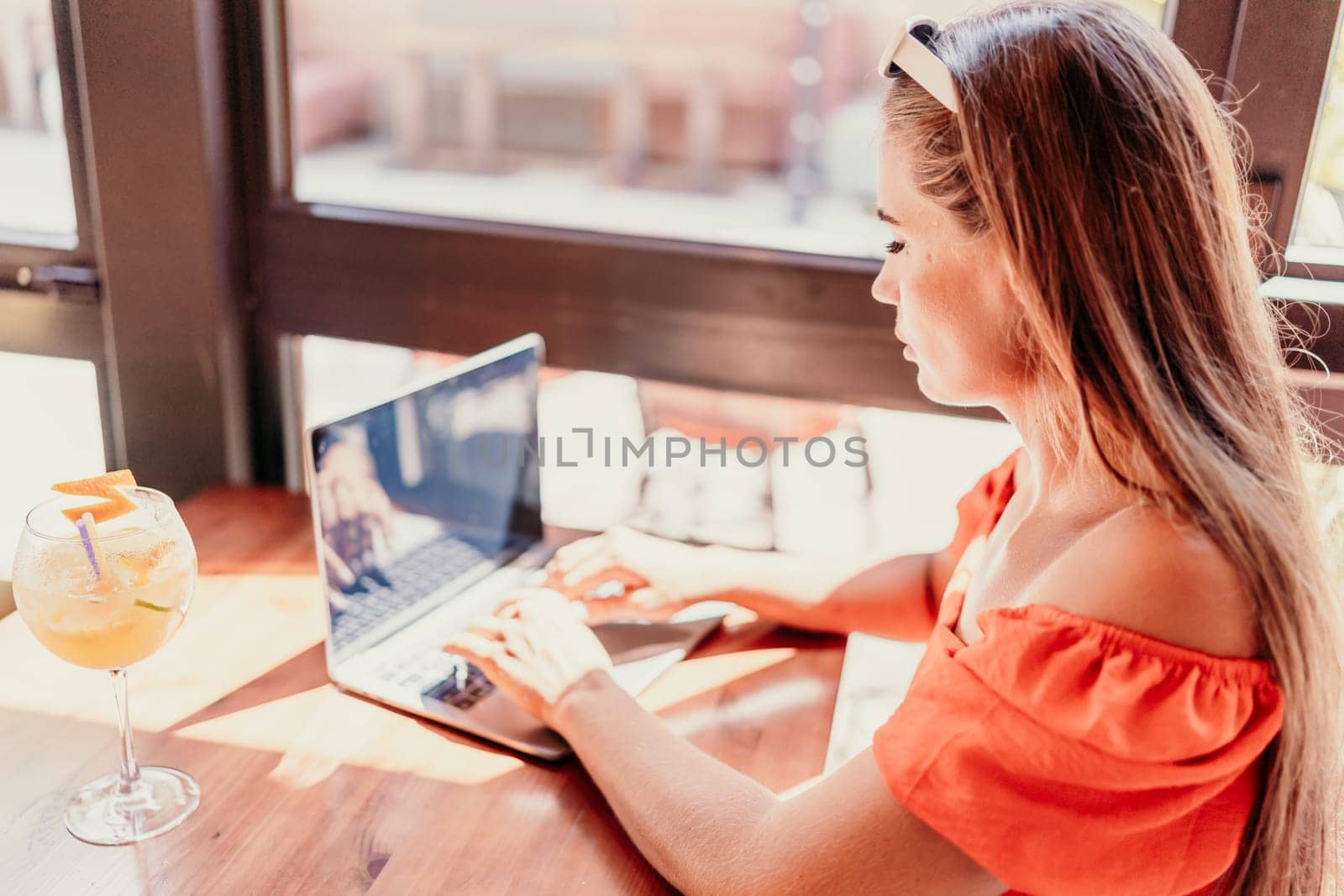 Woman works on laptop in a sunny cafe, glass of juice nearby. The setting is modern, with natural light illuminating the space. The image captures a moment of focused work