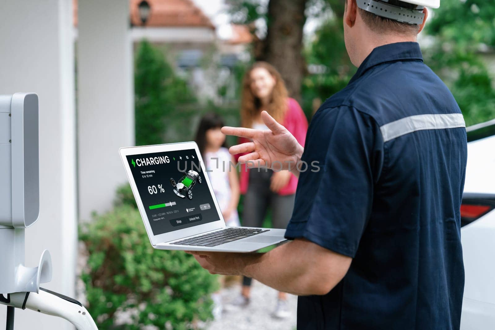 Qualified technician working on home EV charging station installation, making troubleshooting and configuration setup on charging system with laptop for EV at home with the family present. Synchronos