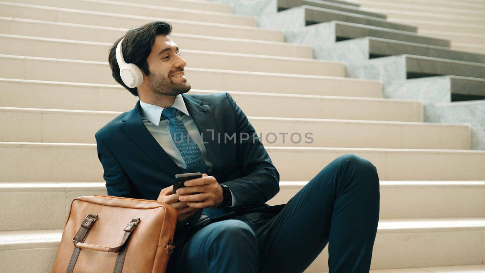 Professional smart business man listening music while lie on stairs. Smart manager happy while receive good news or getting project from headphone. Investor wear suit and bag while sitting. Exultant.