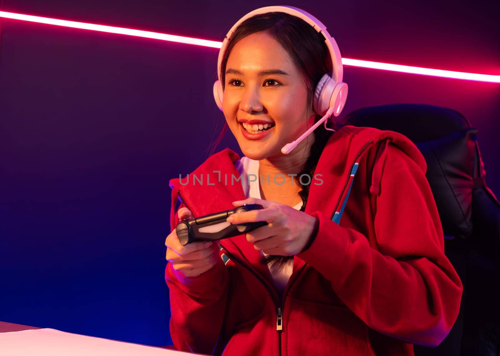 Host channel of smiling beautiful Asian girl streamer with joystick playing online game wearing headphones paste talking with viewers media online. Esport skilled team players in neon room. Stratagem.