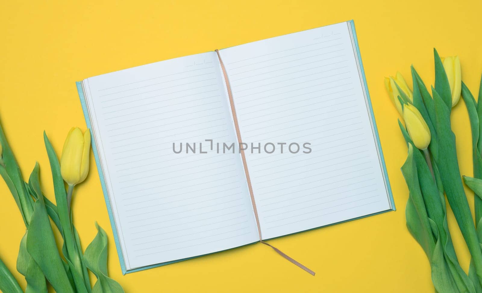 Open notebook with white blank sheets and a bouquet of yellow tulips on a yellow background, top view