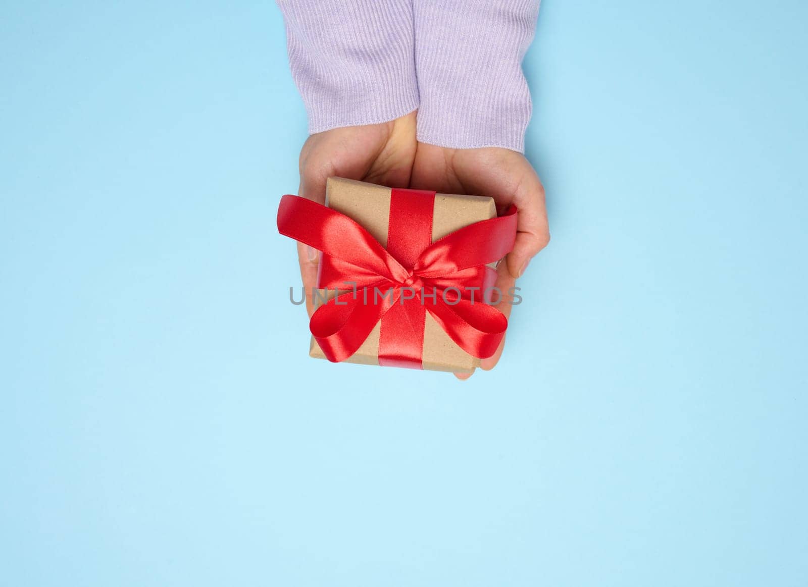 Woman's hand holding a gift box wrapped in a silk ribbon on a blue background, top view