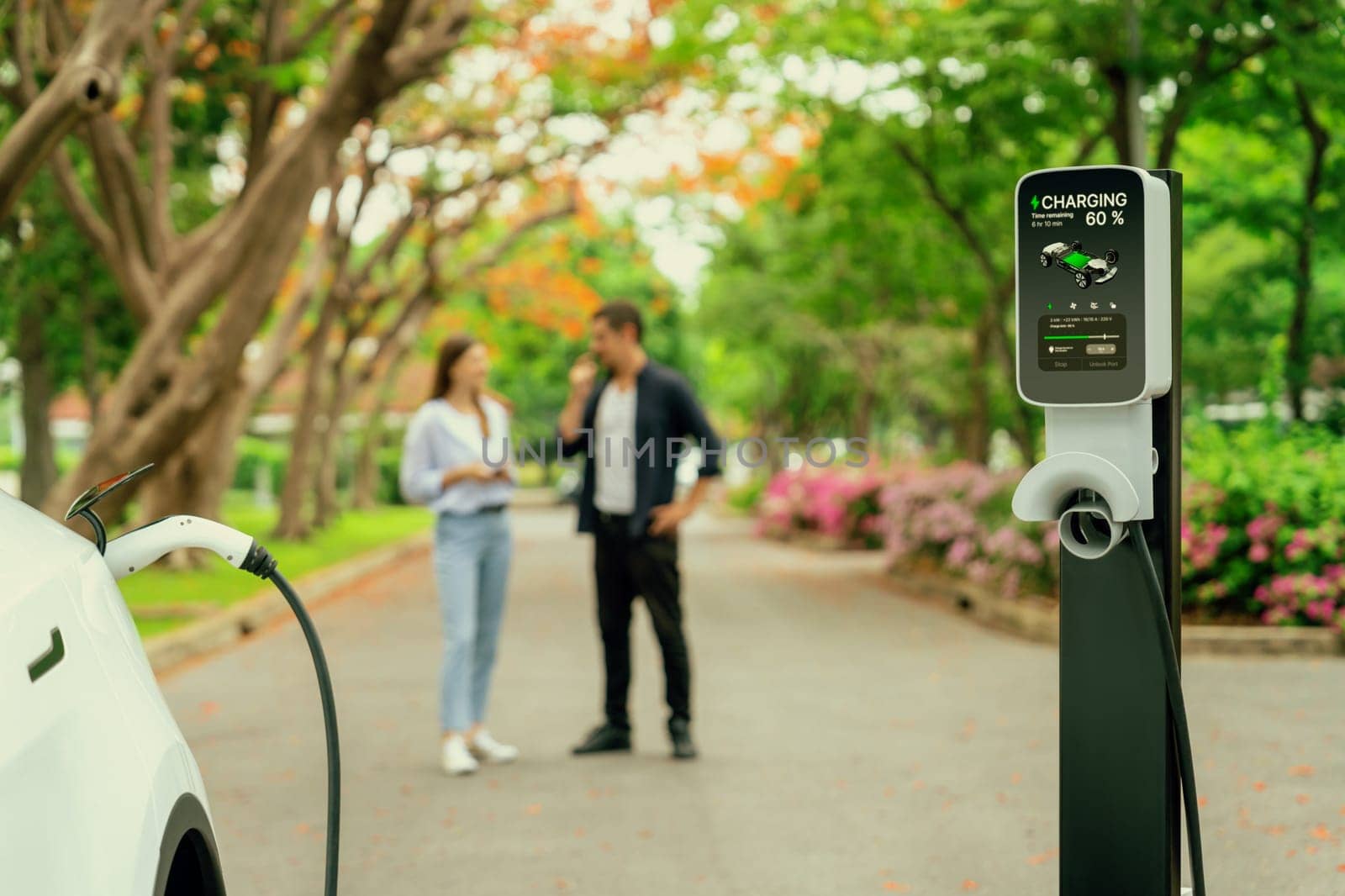 Focused EV car recharging battery on blurred background of lovey couple during autumnal road trip travel with electric vehicle recharging battery. Eco friendly travel on vacation during autumn. Exalt