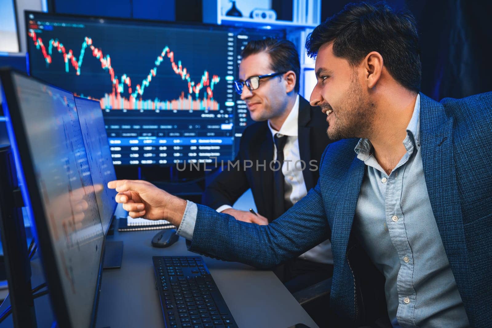 Two stock exchange traders discussing on dynamic successful investment graph, pointing trending of high profit in currency stock in financial data on monitor in neon light at workplace. Sellable.