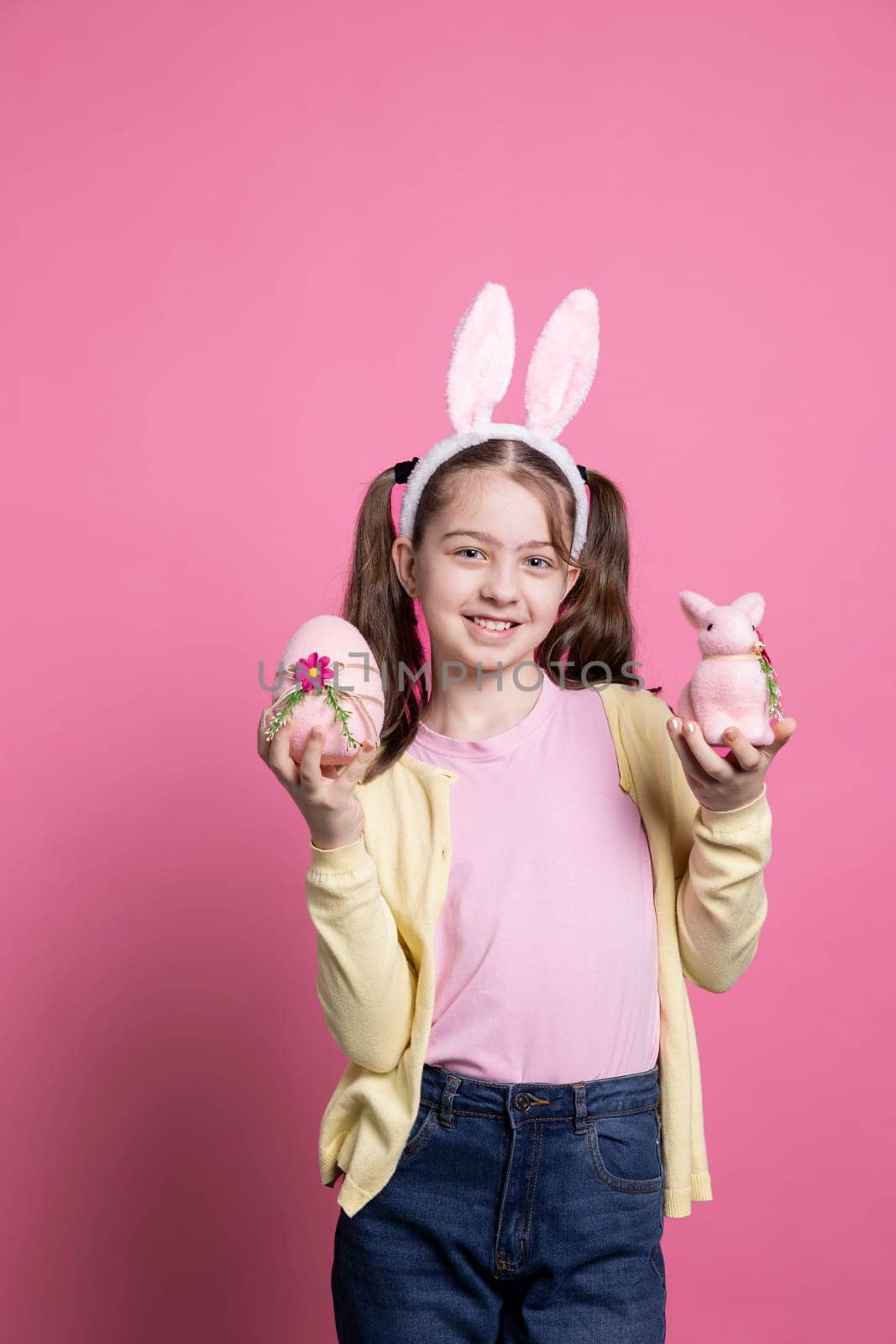 Joyful toddler presenting a pink egg and a stuffed rabbit in studio, wearing pigtails and cute bunny ears on camera. Smiling young kid showing her easter decorations and colorful toys.