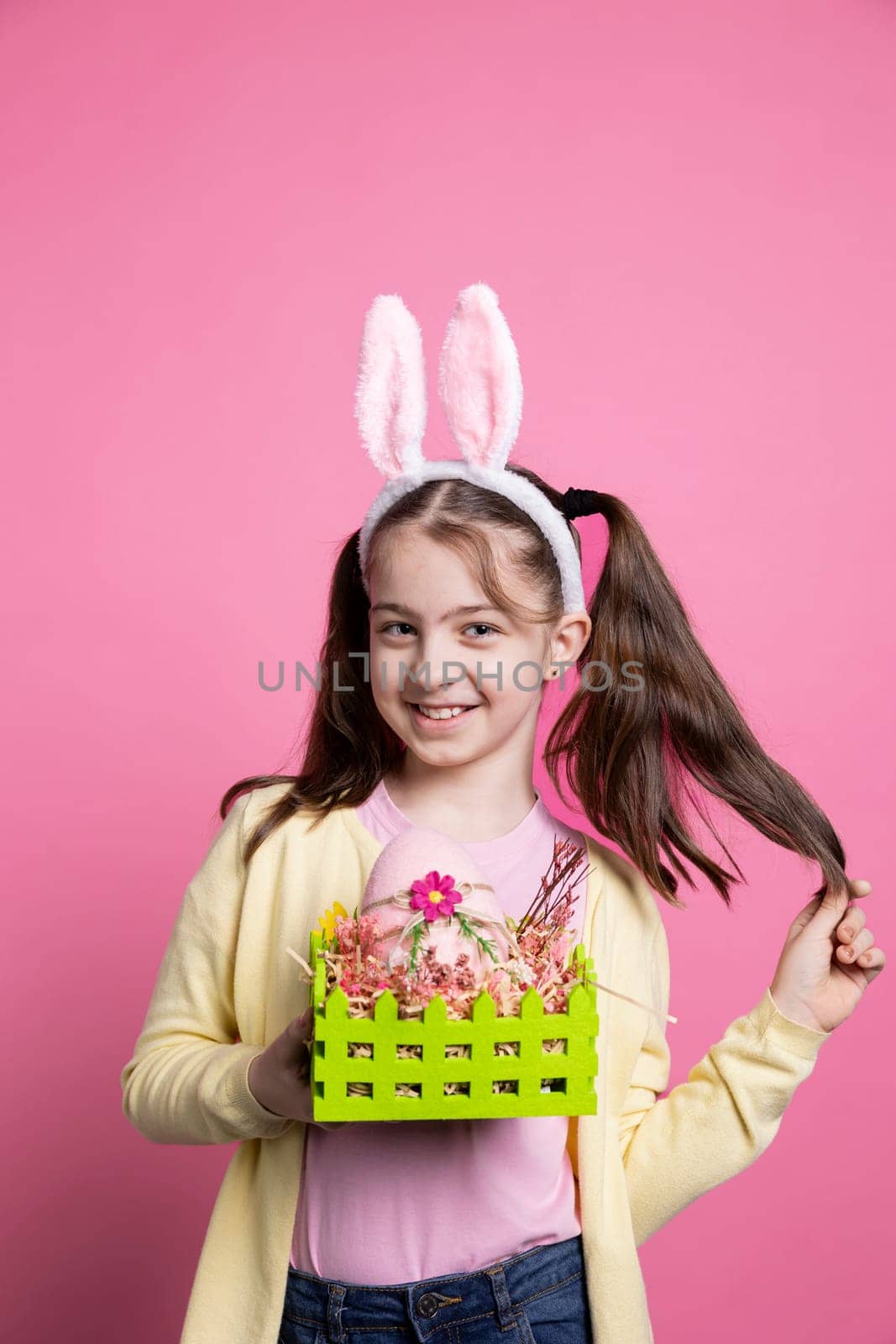 Young cheerful child with bunny ears holding colored basket in studio, posing with painted eggs and ribbons. Small girl excited about easter celebrations, shows handmade ornaments.