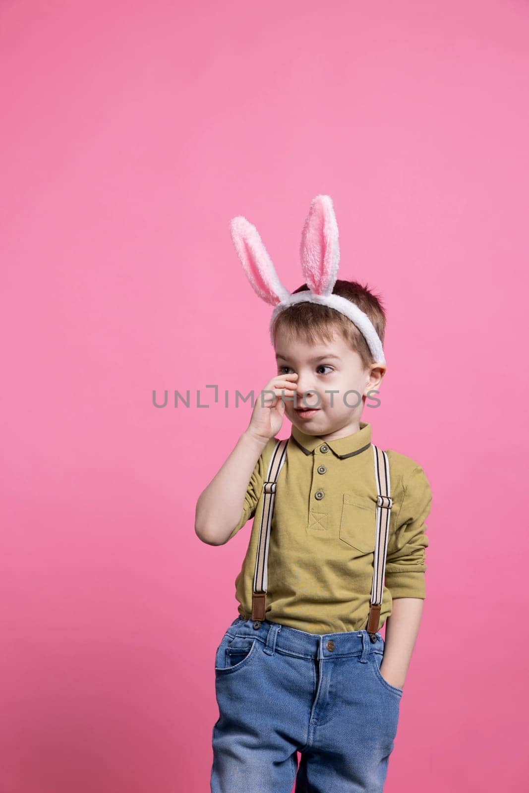 Joyful young kid feeling confident posing against pink background, wearing bunny ears to celebrate easter holiday event. Small little child smiling in front of camera, waiting to receive presents.