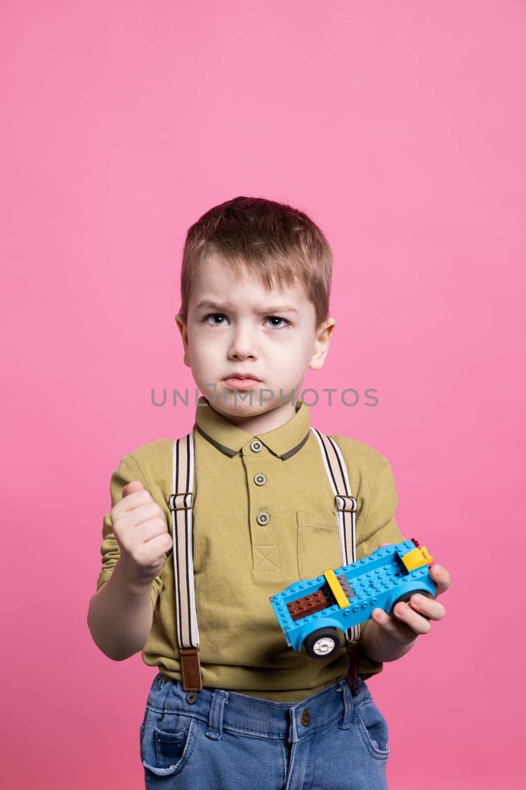 Displeased irritated small boy feeling sad while he plays with a toy car, almost crying in front of the camera. Little child feeling angry and upset holding a plastic blue automobile.