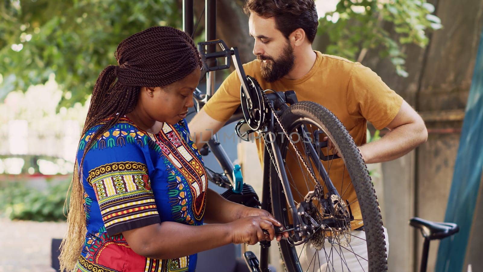 Multiethnic couple working together to repair and maintain damaged bike for summer cycling. Caucasian boyfriend helps girlfriend in choosing suitable tool for fixing bicycle hub and axle in yard.