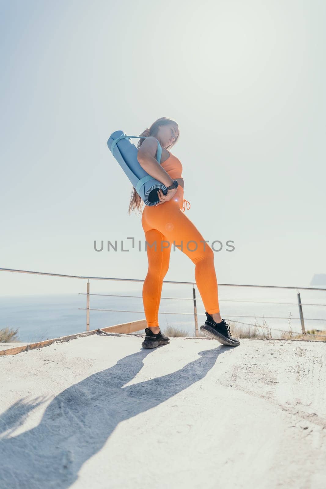 Fitness woman sea. Happy middle aged woman in orange sportswear exercises morning outdoors on yoga mat with laptop in park over ocean beach. Female fitness pilates yoga routine. Healthy lifestyle. by panophotograph
