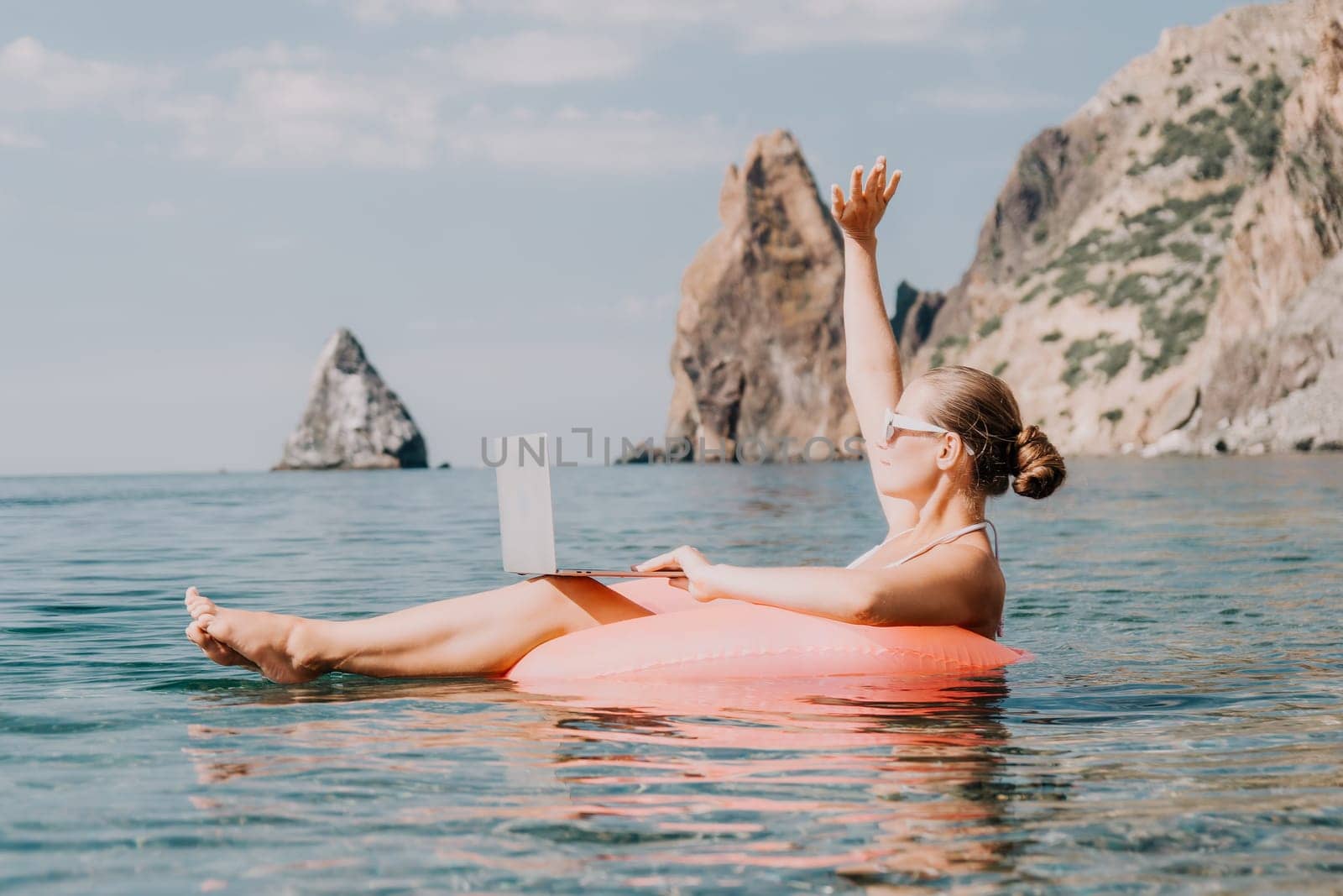 Woman freelancer works on laptop swimming in sea on pink inflatable ring. Pretty lady typing on computer while floating in the sea on inflatable donut at sunset. Freelance, remote work on vacation