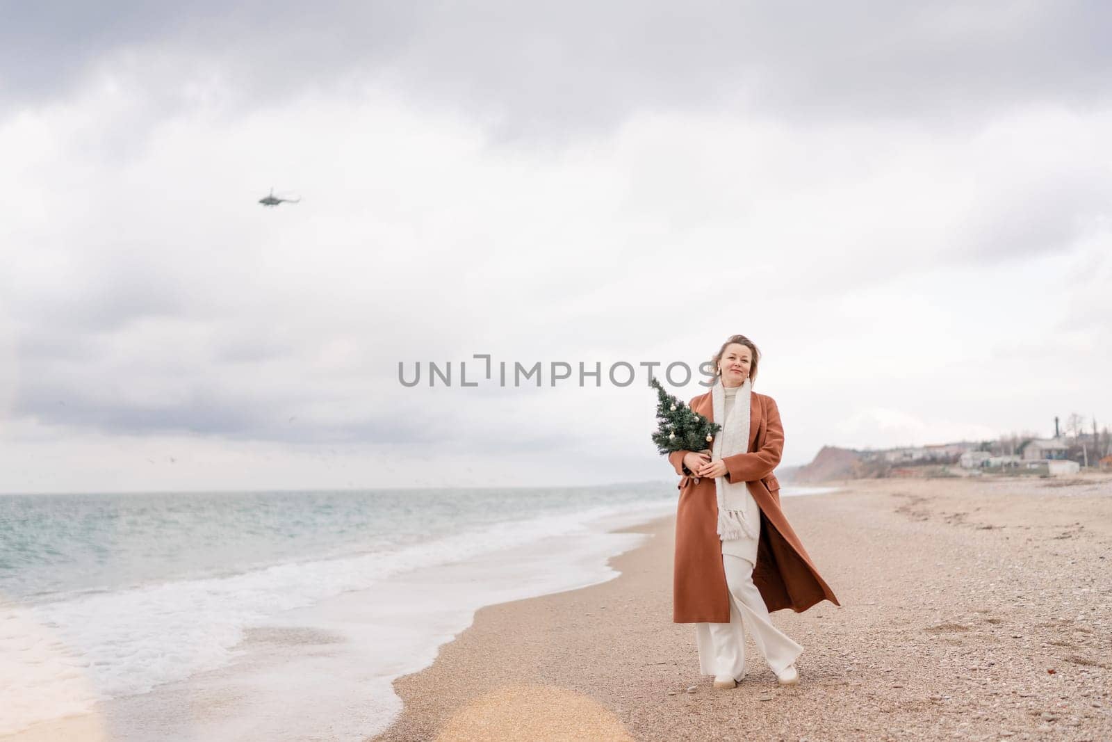 Blond woman Christmas sea. Christmas portrait of a happy woman walking along the beach and holding a Christmas tree in her hands. She is wearing a brown coat and a white suit