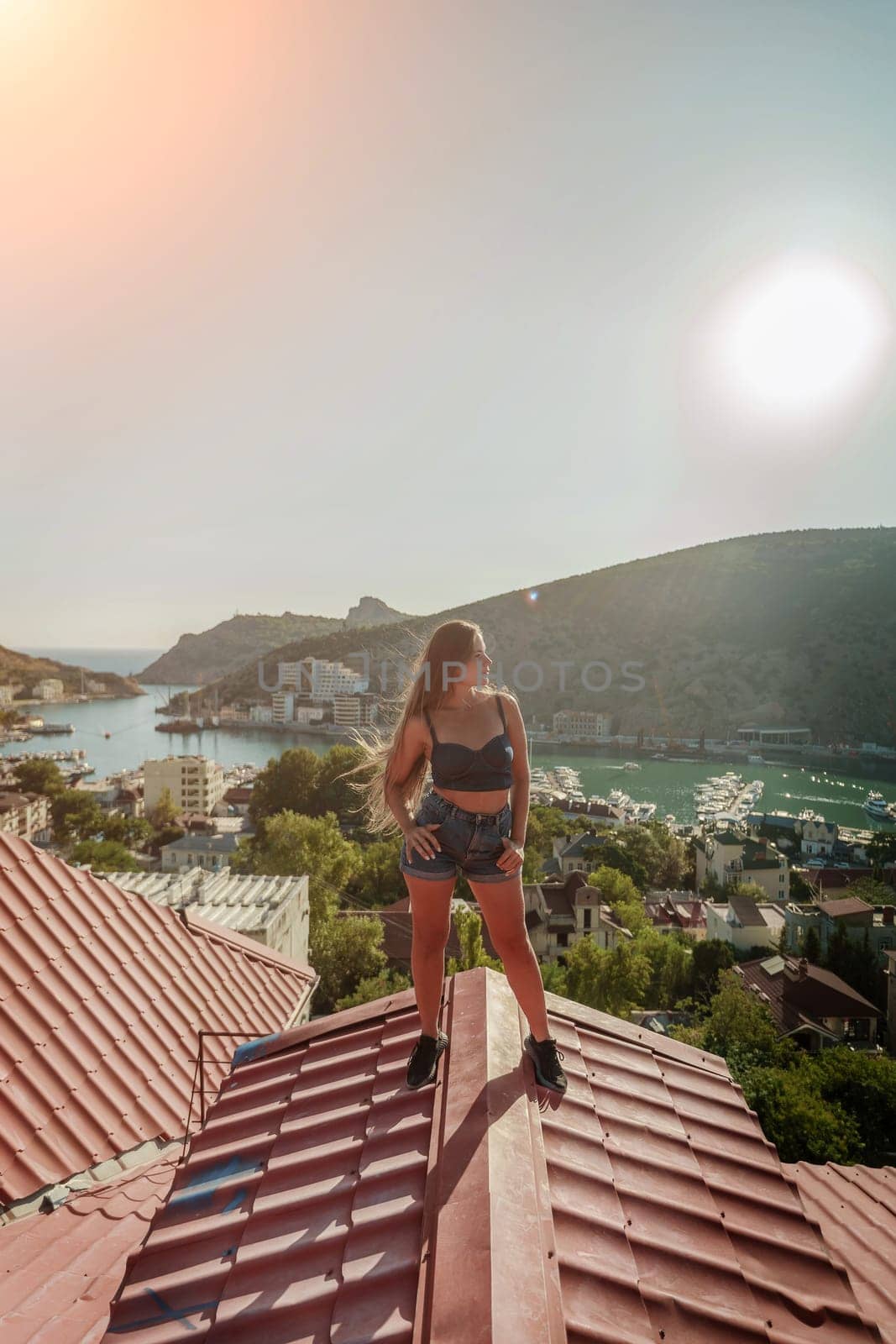 Woman standing on rooftop, enjoys town view and sea mountains. Peaceful rooftop relaxation. Below her, there is a town with several boats visible in the water. Rooftop vantage point