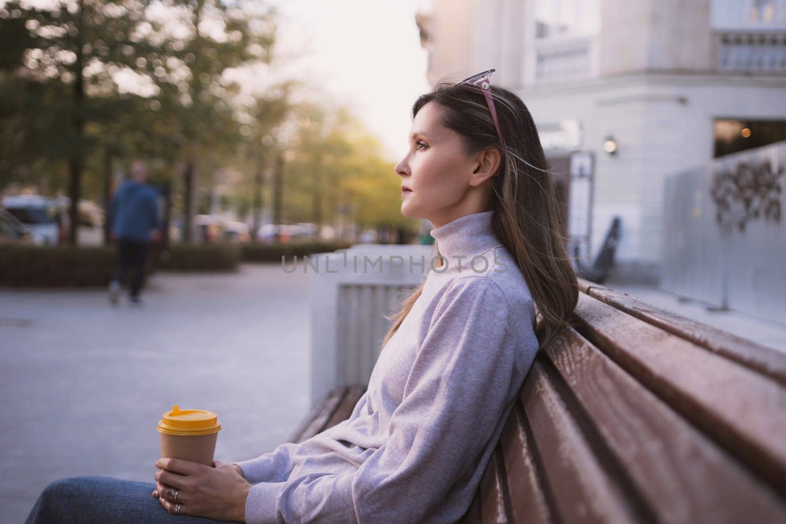 Woman drinks from cup on wooden bench. She is wearing a white shirt enjoying her beverage. The bench is located in a park setting, with trees in the background. by Matiunina