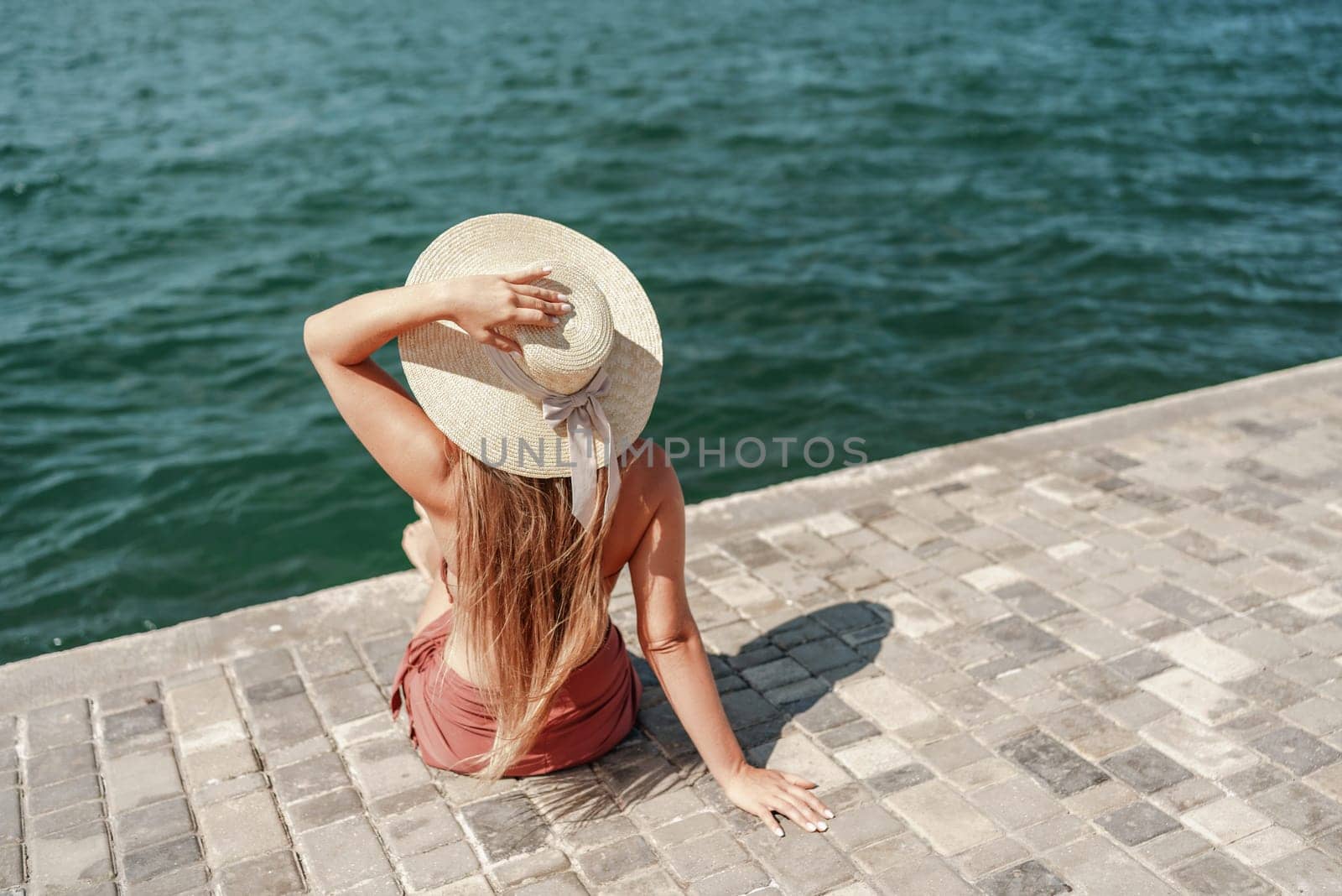 A woman in a swimsuit sits with her back holding a hat, looks at the ocean, sunny day, relaxes