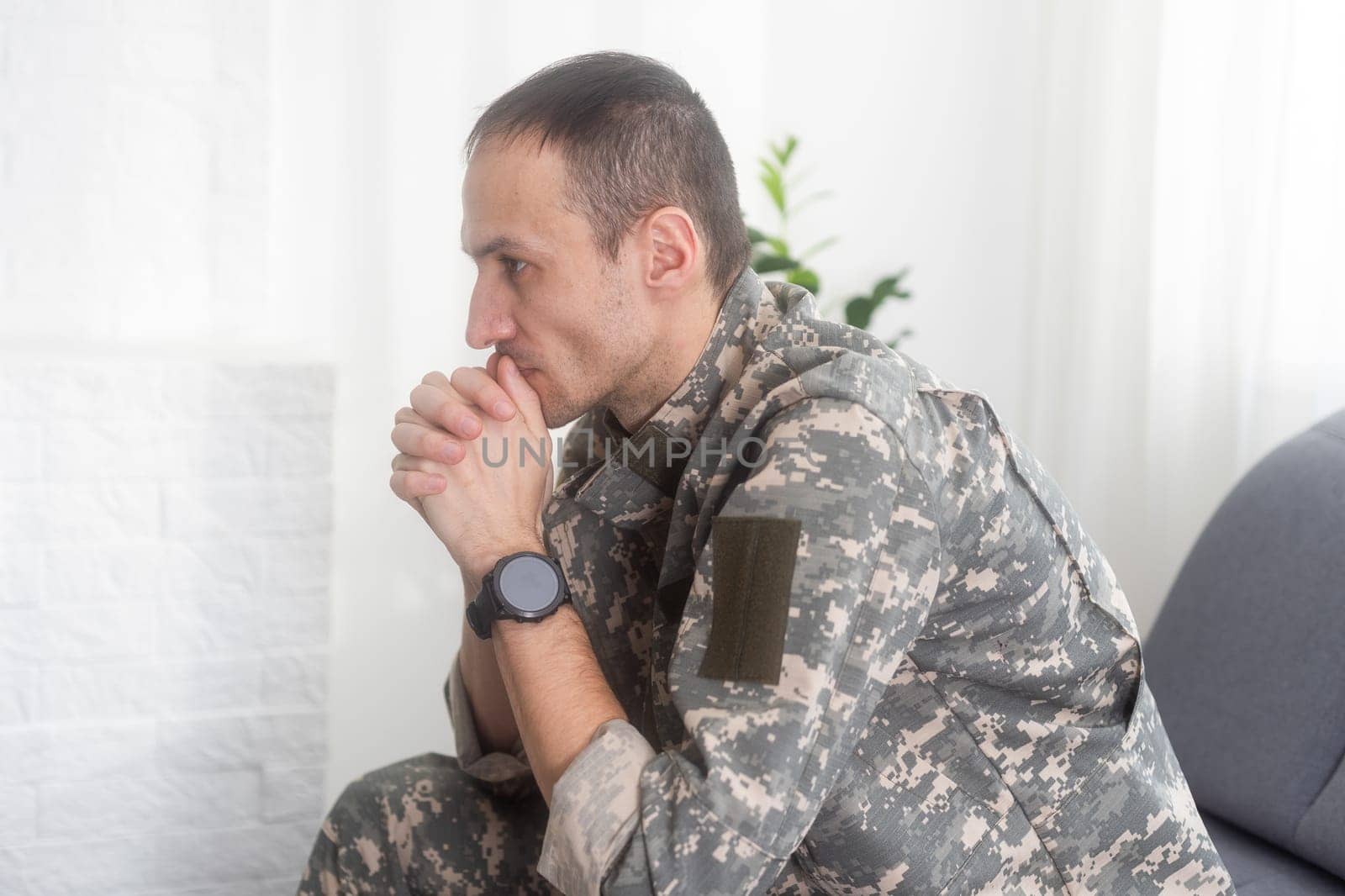 Portrait of calm serious Caucasian military man wearing camouflage uniform and cap sitting on a sofa, having depressed facial expression. High quality photo
