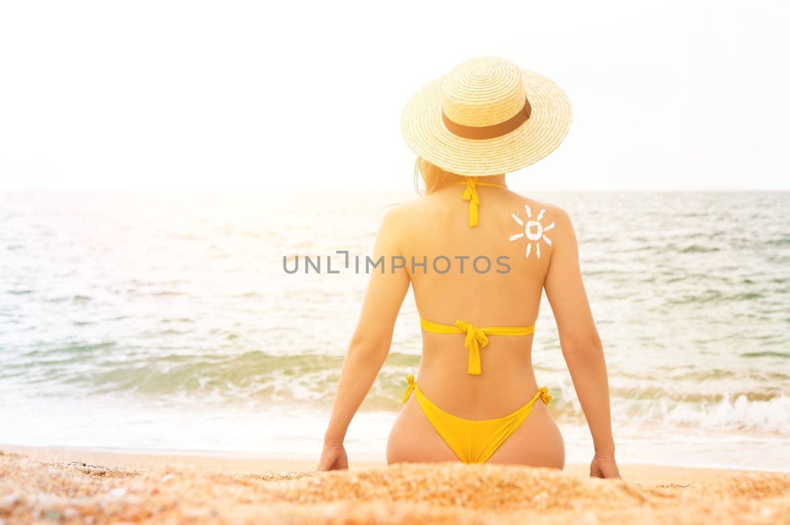 A young Caucasian woman in a swimsuit and a straw hat sits on the beach in front of the sea with her back in the frame. The sun on a woman's back is drawn with sunscreen.