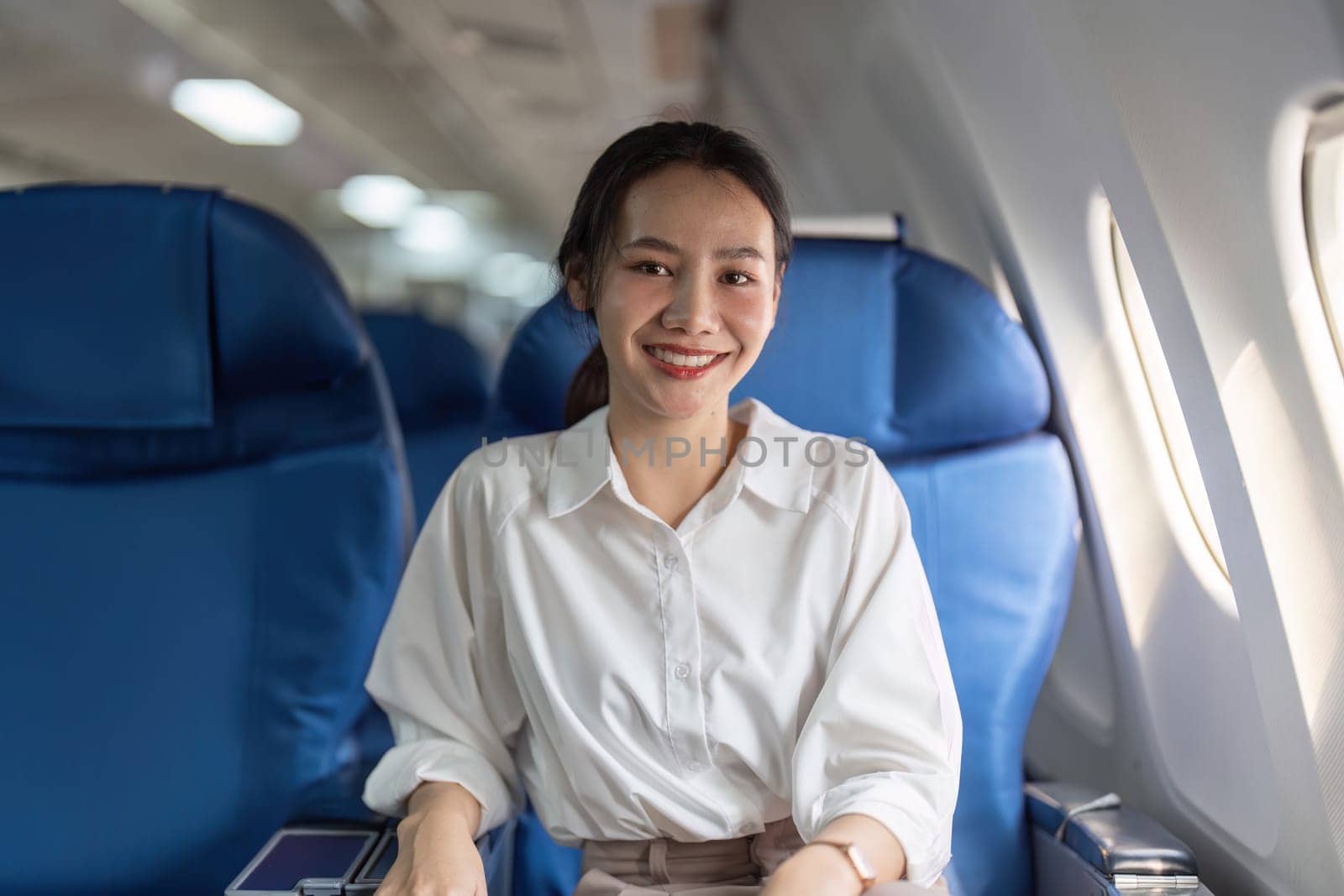 A woman is sitting on a blue airplane seat with a smile on her face. She is wearing a white shirt and a ponytail