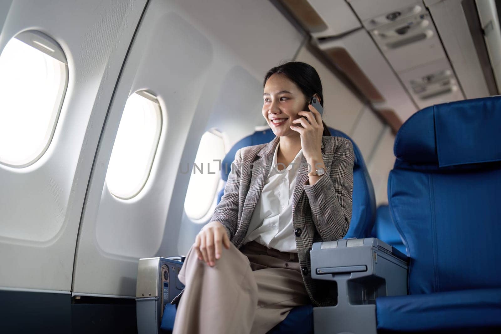 Asian young businesswoman successful or female entrepreneur in formal suit in a plane sit in a business class seat and uses a smartphone during flight. Traveling and Business concept.