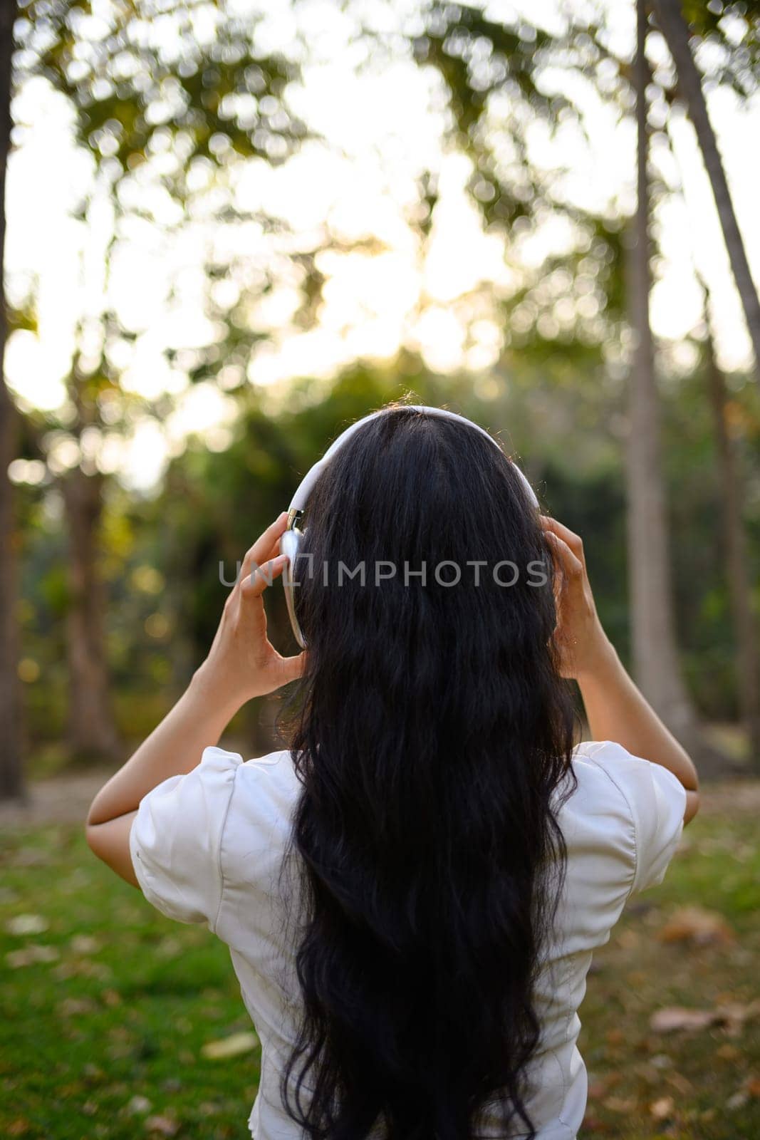 Back view of young woman standing alone in the park and listening music with headphones.