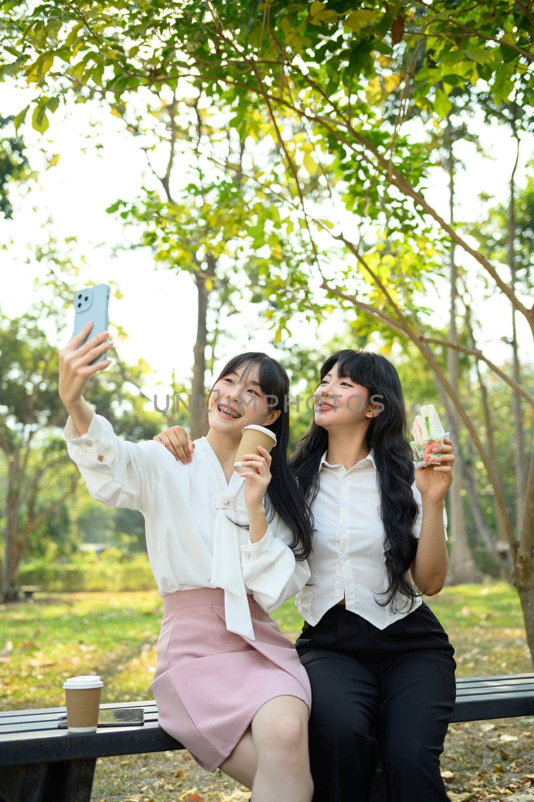 Cheerful Asian female friends sitting on park bench and taking a selfie during lunch break.
