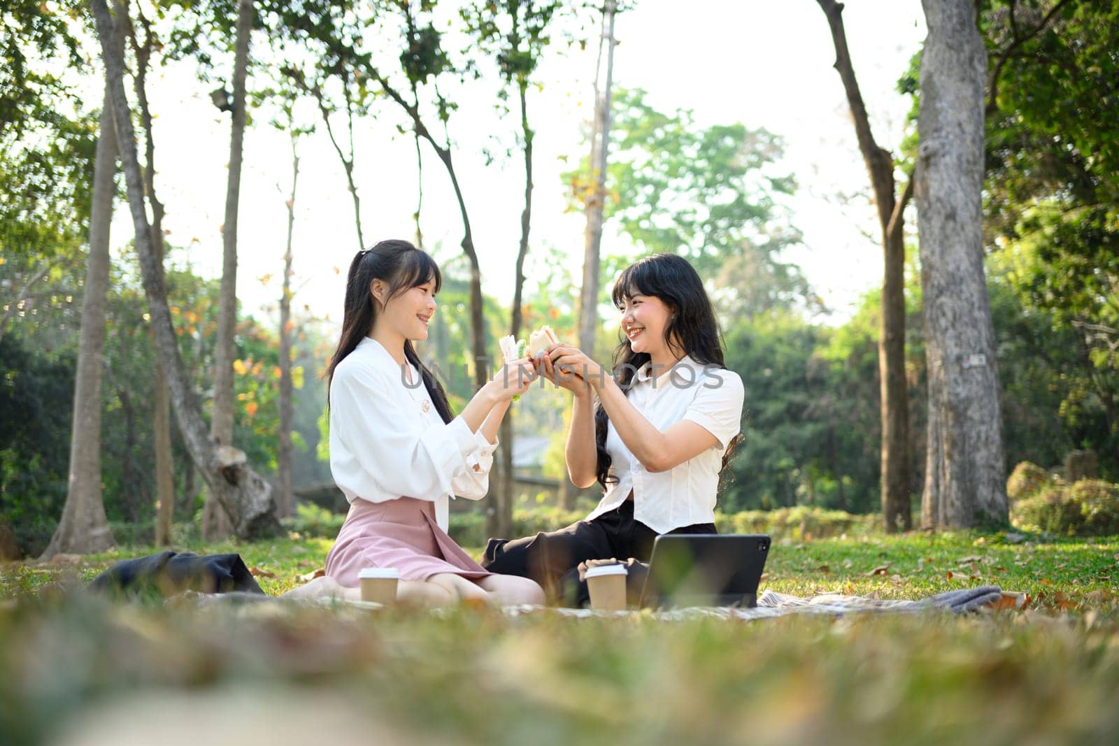 Young woman friends sitting on grass and eating sandwiches during picnic in park