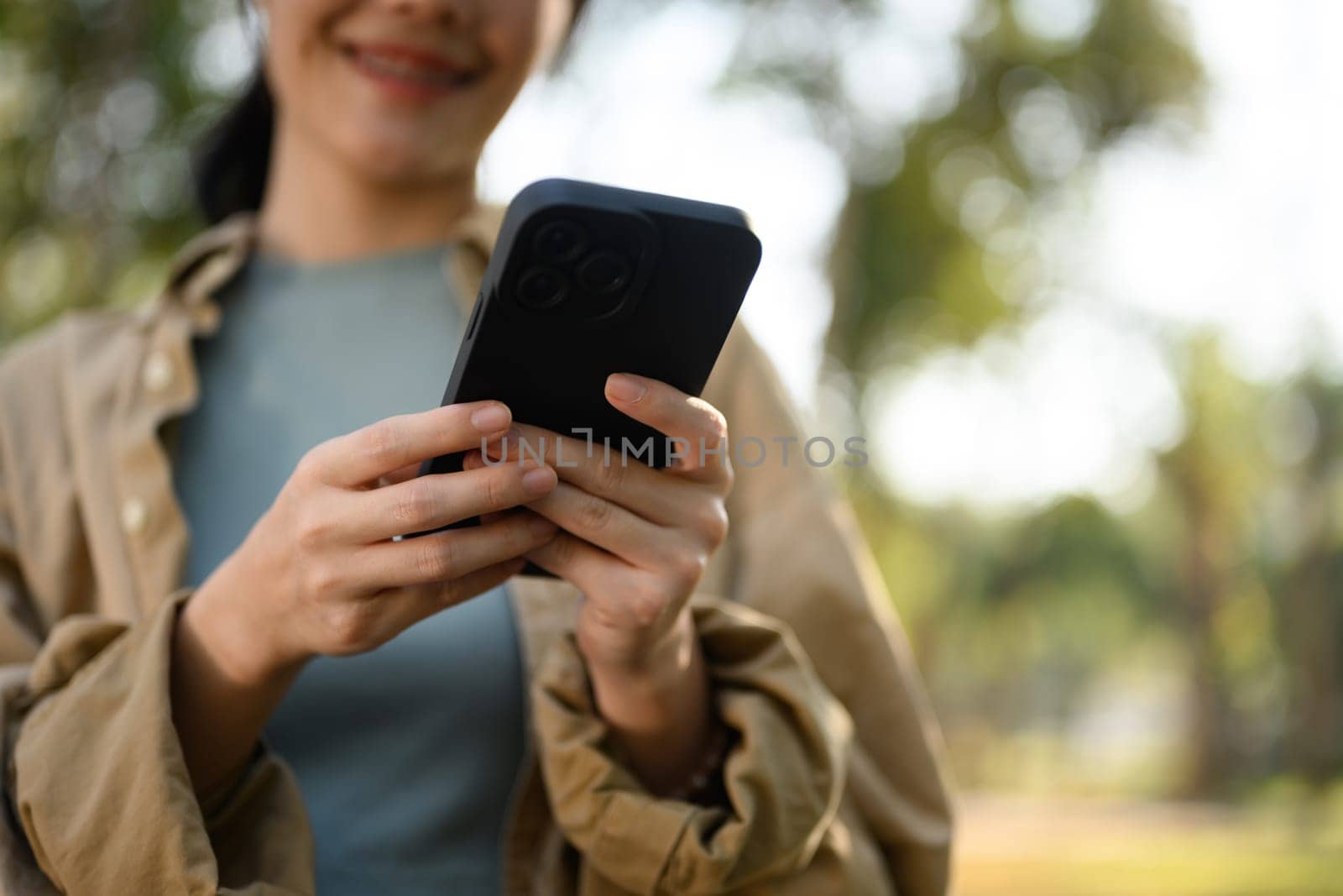 Smiling young lady reading text message, typing on mobile phone while sitting in the park by prathanchorruangsak