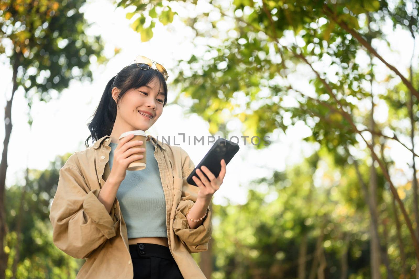Smiling young lady sitting in the park drinking coffee and checking her mobile phone.