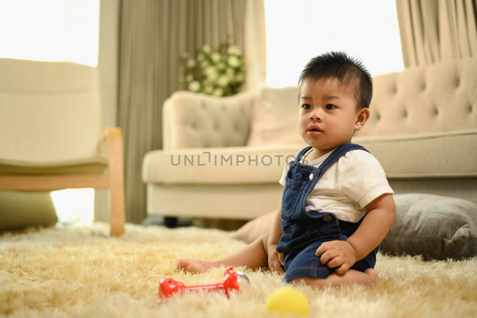 Cute little baby boy sitting on fluffy carpet on and playing with toys in living room.