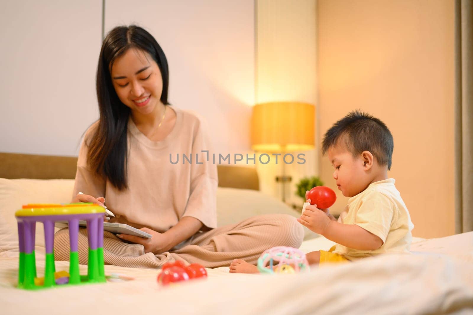 Cute little baby boy playing with toys near working mom in bedroom. Motherhood and child care concept by prathanchorruangsak