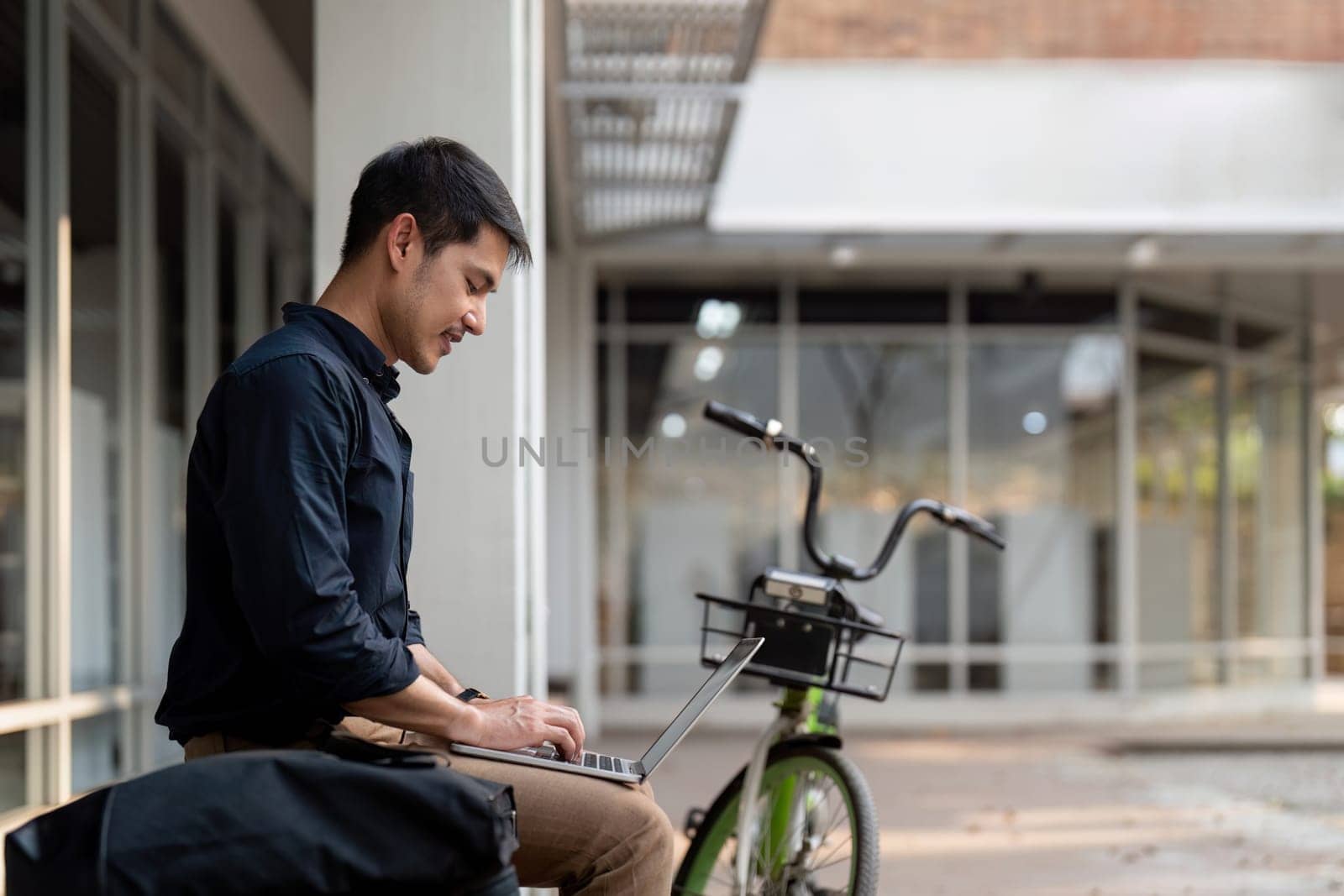 Asian businessman with bicycle using laptop and sitting outside the office building. man commuting on bike go to work. Eco friendly vehicle, sustainable lifestyle concept by nateemee